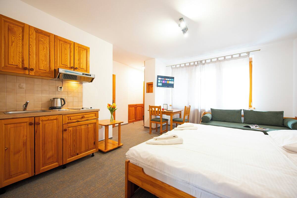 Habitación de hotel con cama, un pequeño escritorio con silla, kitchenette con armarios de madera y televisor en la pared.