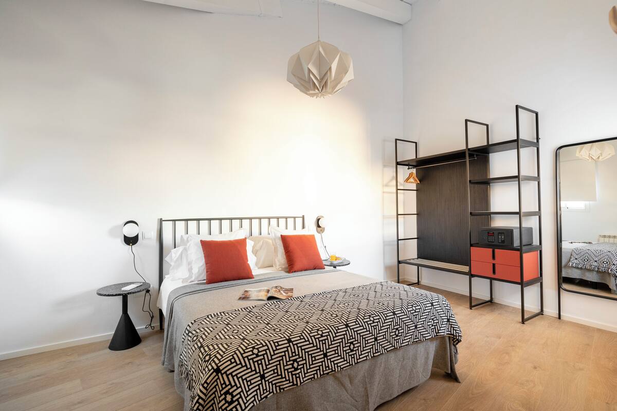 Een moderne slaapkamer met een bed, nachtkastje, lamp, kast en spiegel.