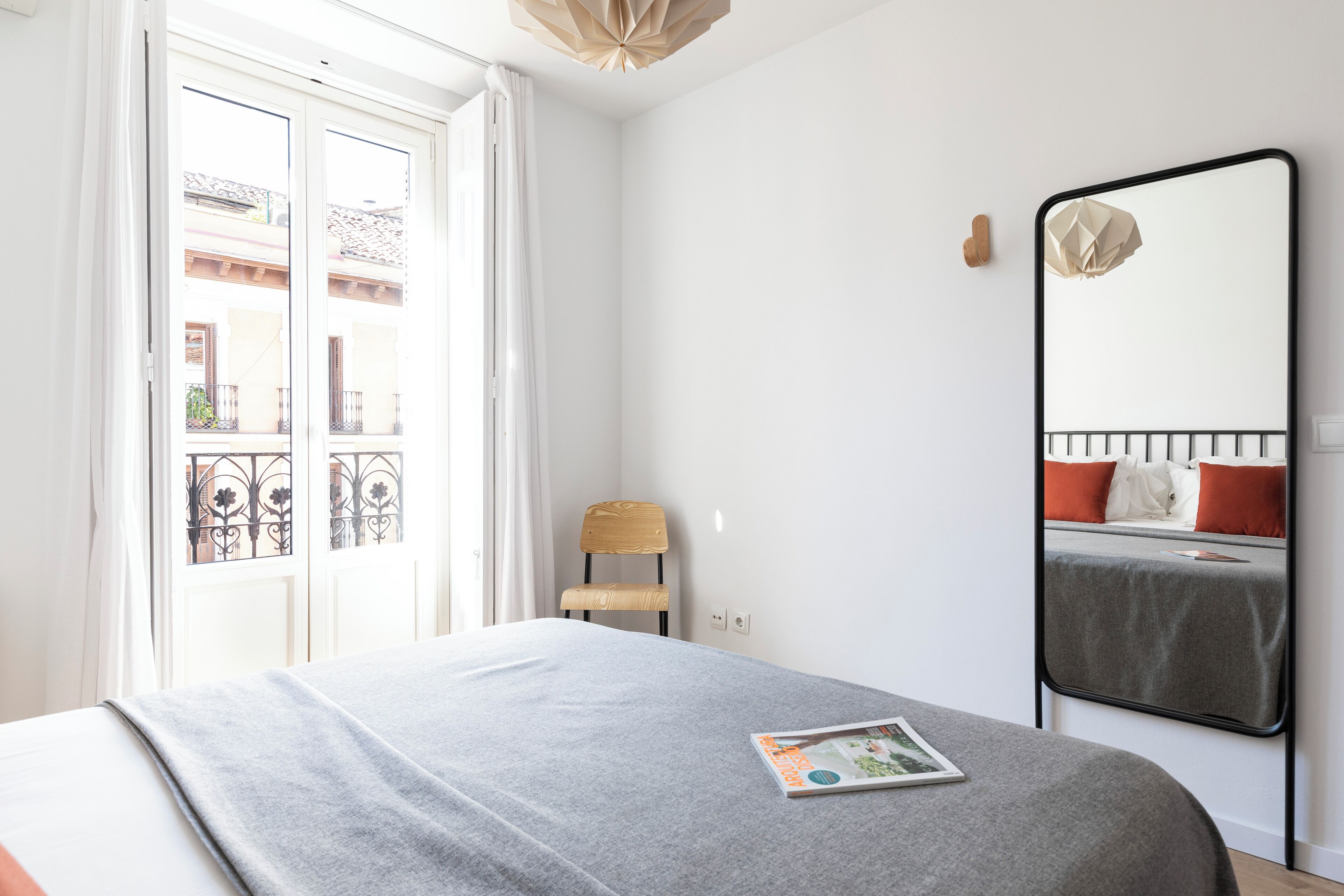 A bedroom with a bed, a chair, a large mirror, and a window with a view of buildings.