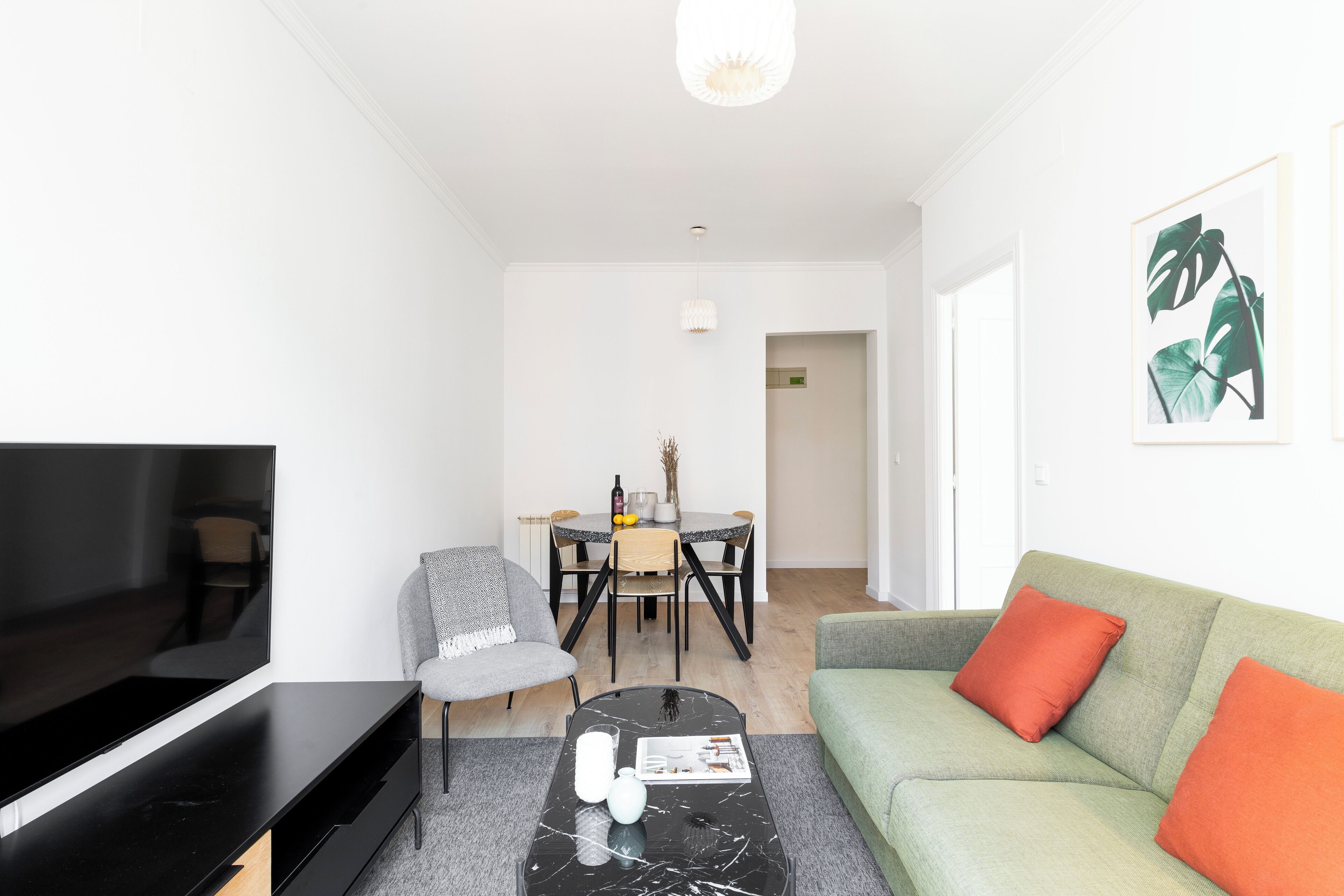 Una sala de estar moderna con televisor de pantalla plana, una mesa de centro negra, un sillón gris, un sofá verde con cojines naranjas, y un comedor con mesa y sillas.