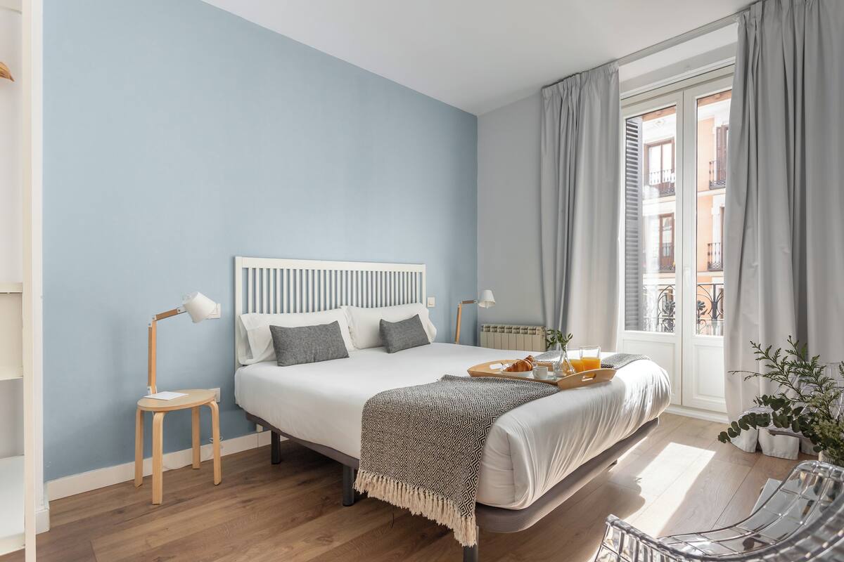 Een slaapkamer met een bed, nachtkastjes, een raam met gordijnen en uitzicht op gebouwen.