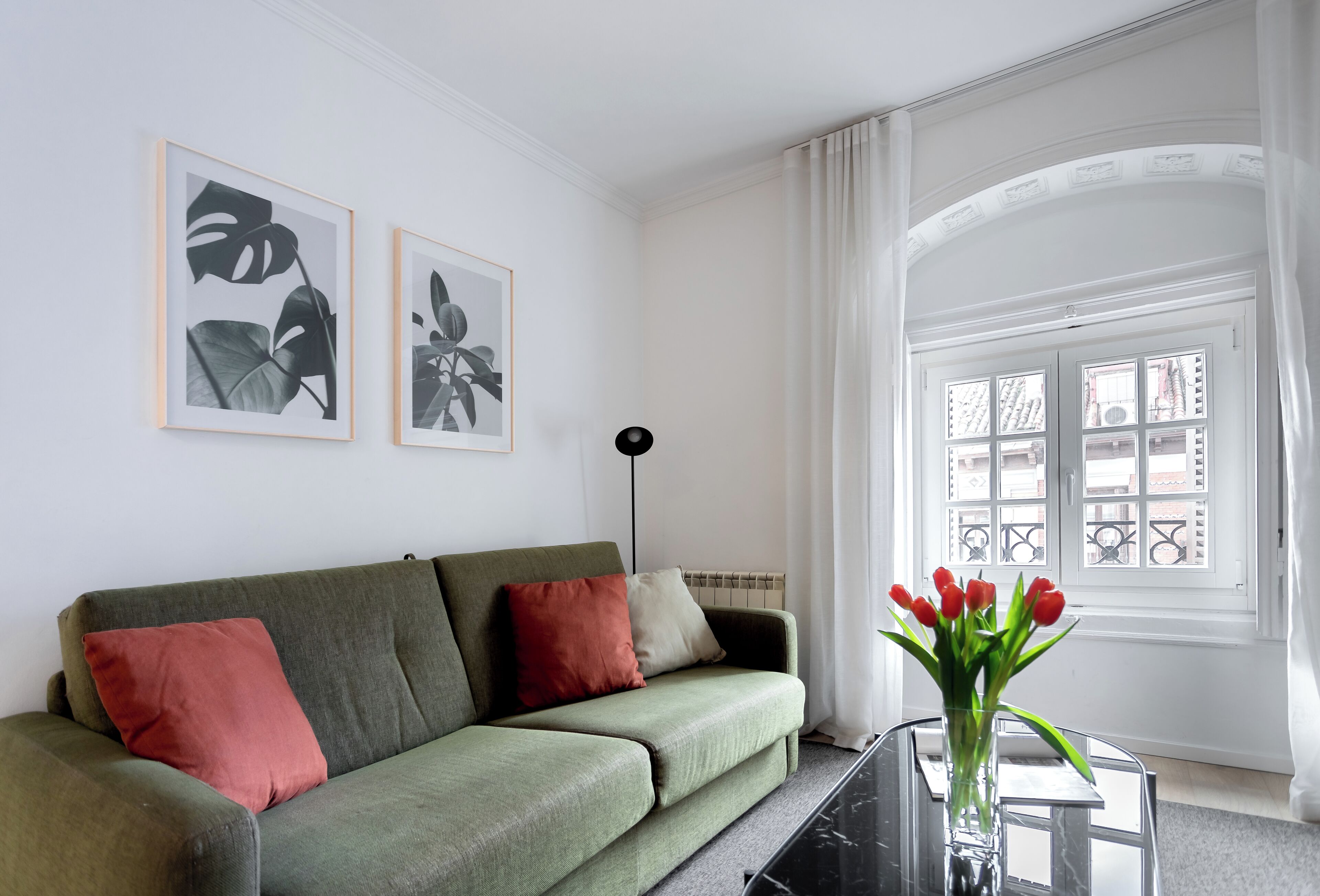 A living room with a green sofa, two framed botanical prints, a vase with red tulips, and a window with a view of buildings.