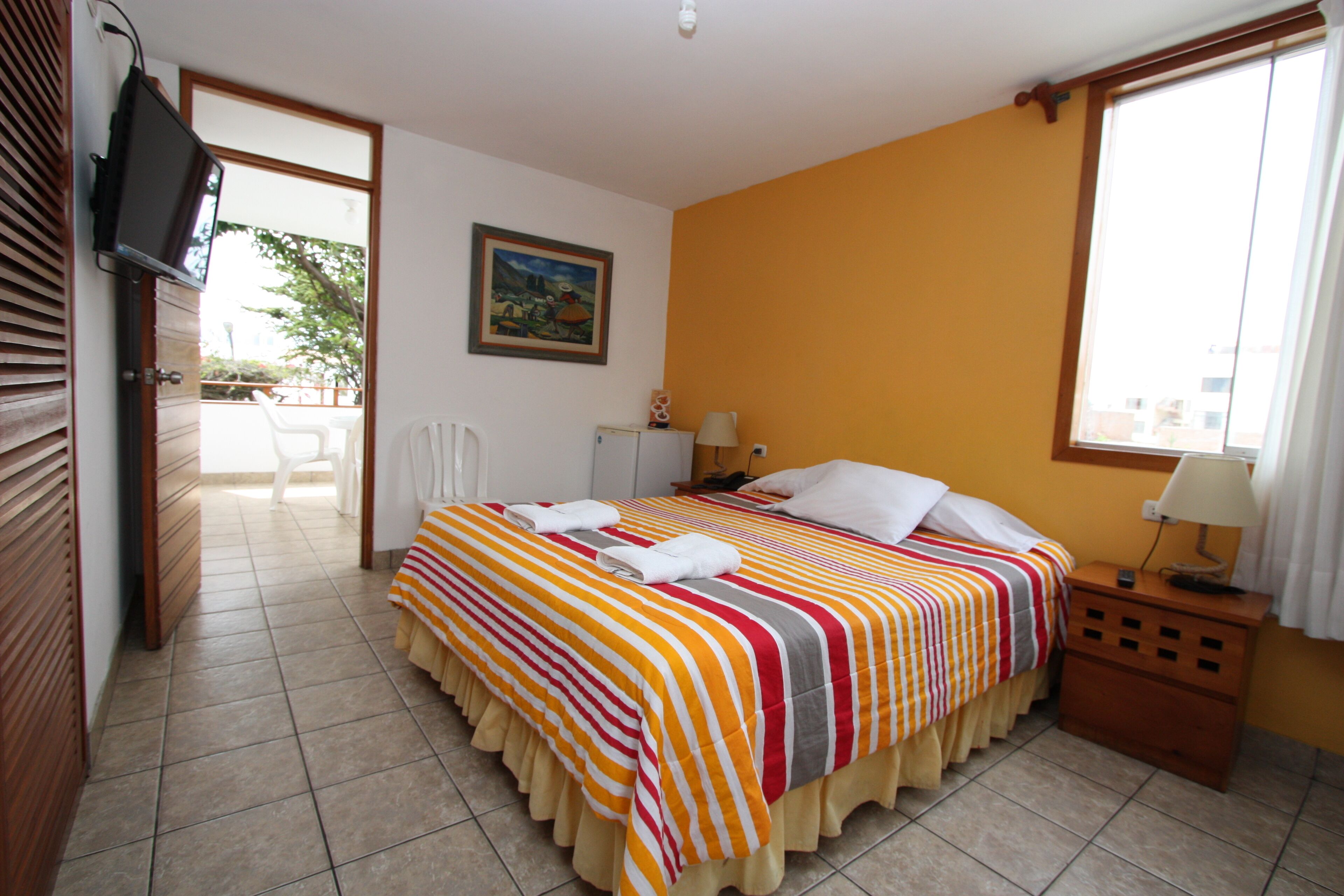 Habitación de hotel con cama, televisión, ventana y puerta que da a un balcón.