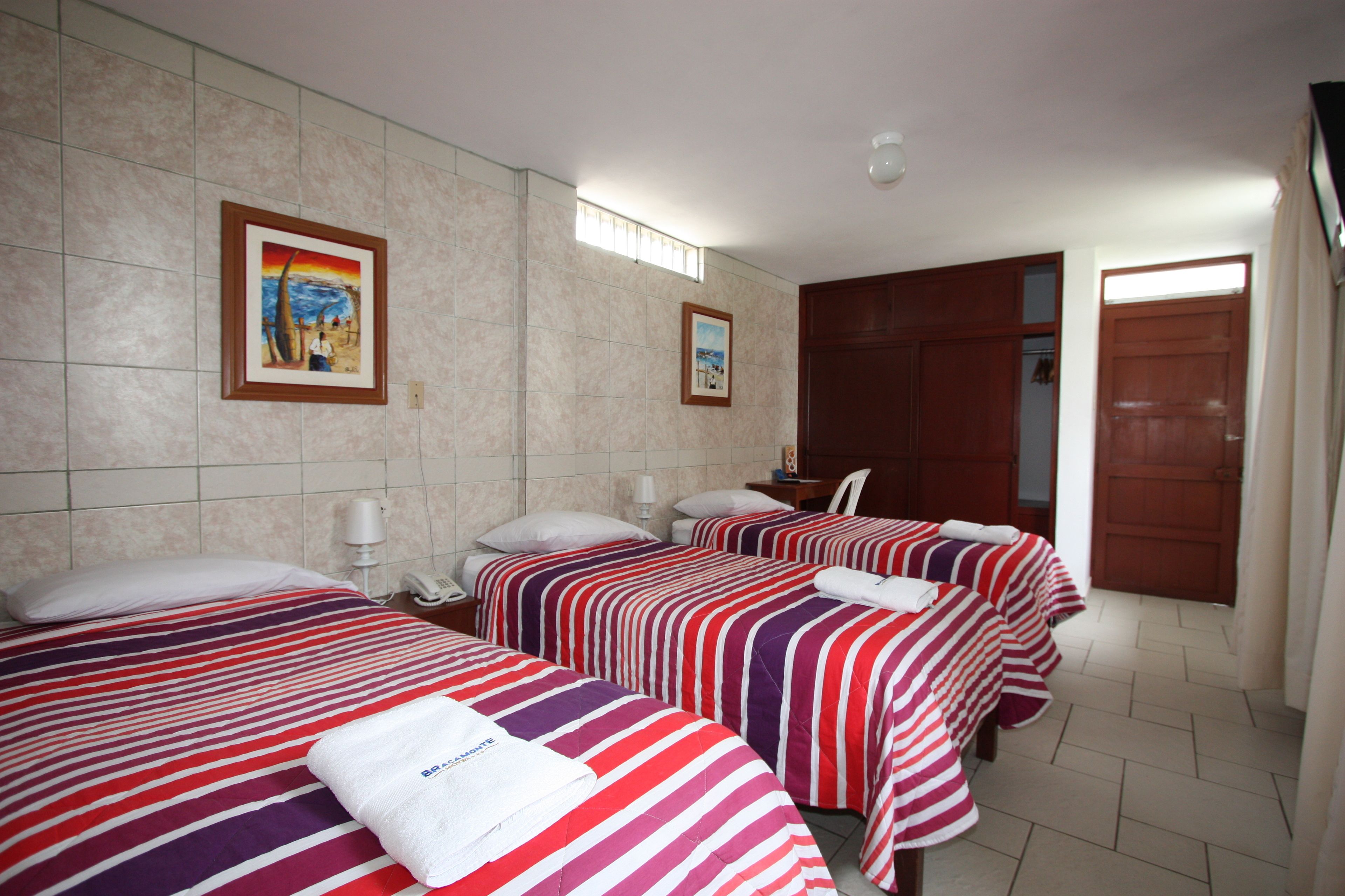 Habitación de hotel con tres camas, cada una con ropa de cama a rayas, un escritorio pequeño con una lámpara y dos cuadros en la pared.