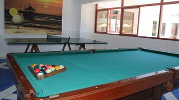 A pool table with balls, a glass table, and a sunset painting on the wall.
