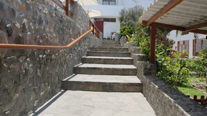 A stone staircase leading to a white building with a red door, surrounded by greenery and a covered area with a wooden railing. - TURISMO BRACAMONTE SAC (Huanchaco)