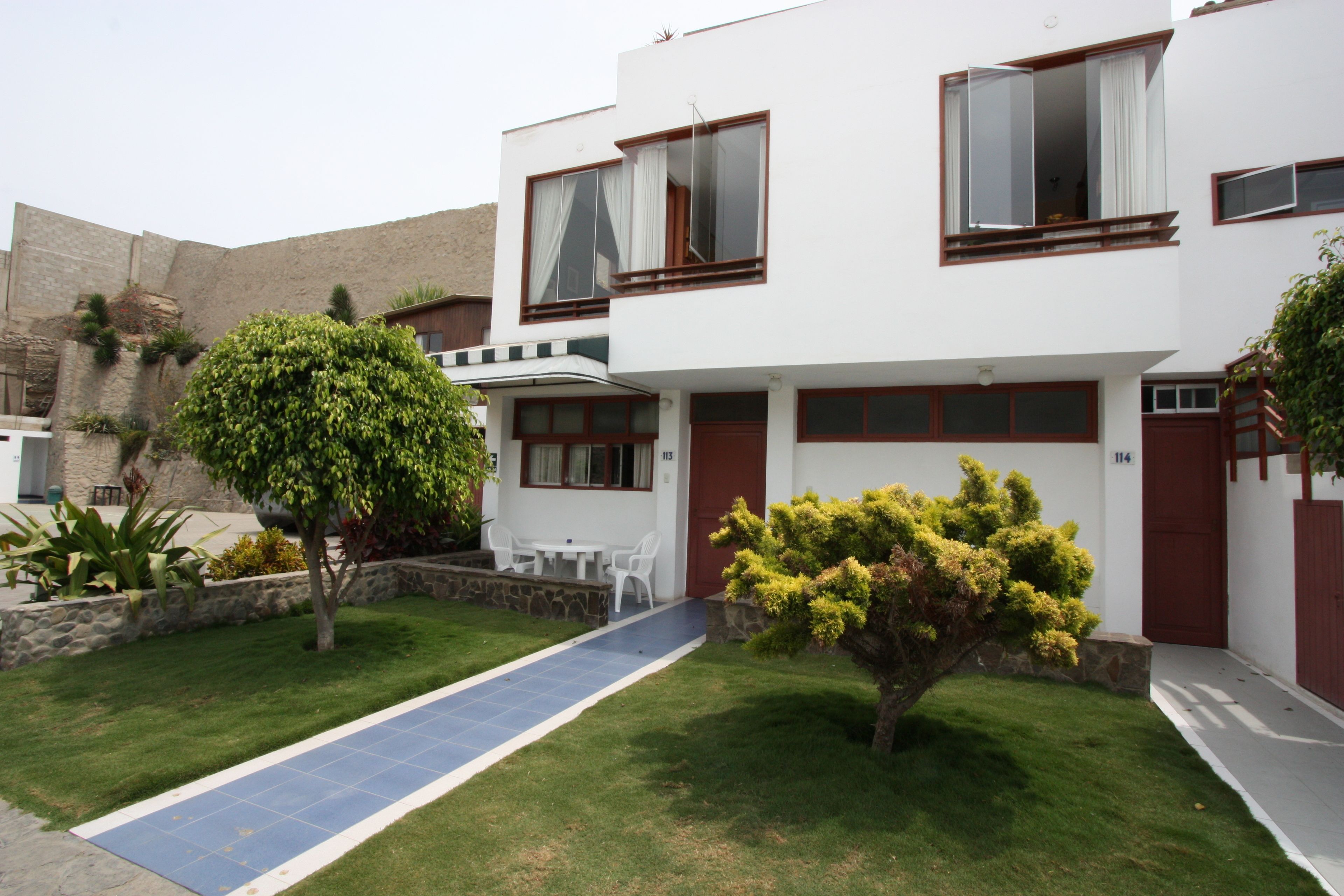 Une maison moderne à deux étages, dotée d’une allée pavée, d’un petit patio avec une table et des chaises, et d’une pelouse bien entretenue.