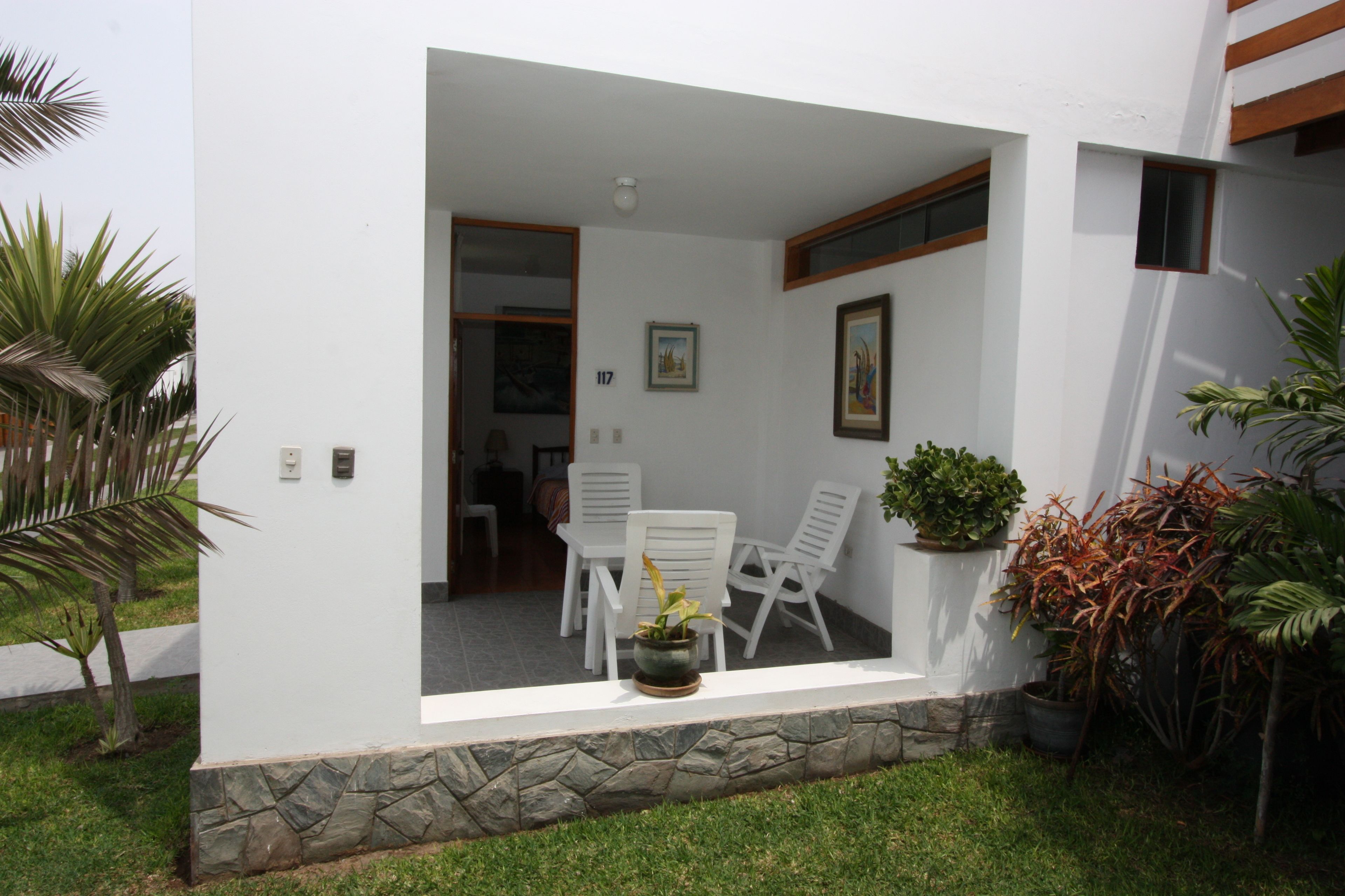 Un patio moderno al aire libre con base de piedra, paredes blancas y una puerta que da a un espacio interior. Hay sillas blancas y una planta en maceta en el patio.