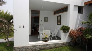 Un patio moderno al aire libre con base de piedra, paredes blancas y una puerta que da a un espacio interior. Hay sillas blancas y una planta en maceta en el patio.