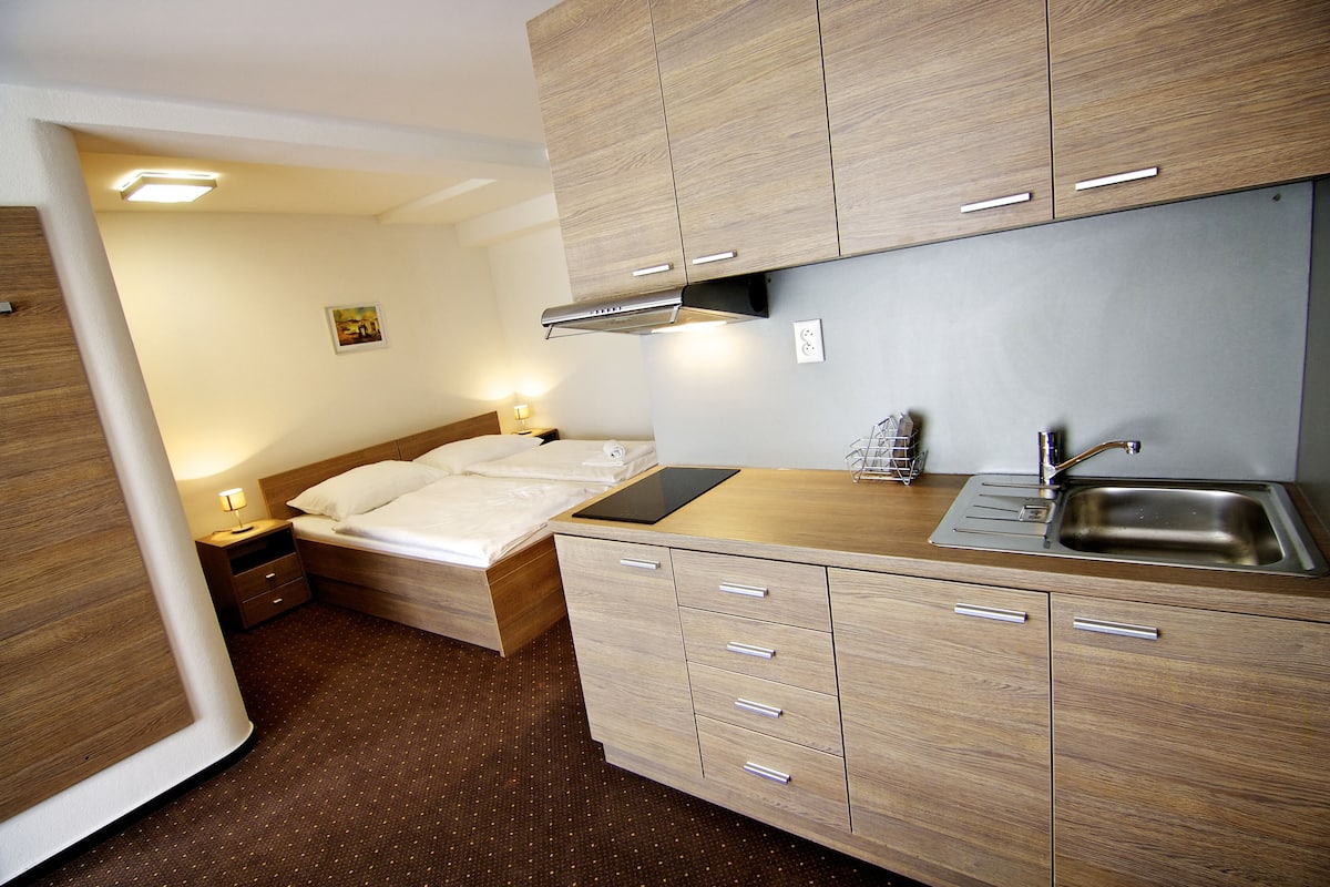 Quarto de hotel compacto com cama, mesa de cabeceira e área de cozinha compacta, incluindo pia e armários.