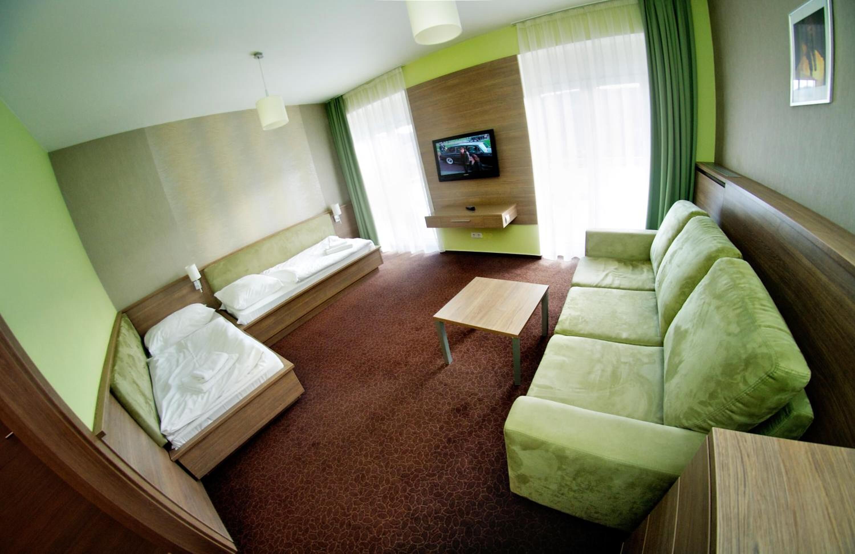 A hotel room with two single beds, a sofa, a small table, a TV mounted on the wall, and a window with curtains.
