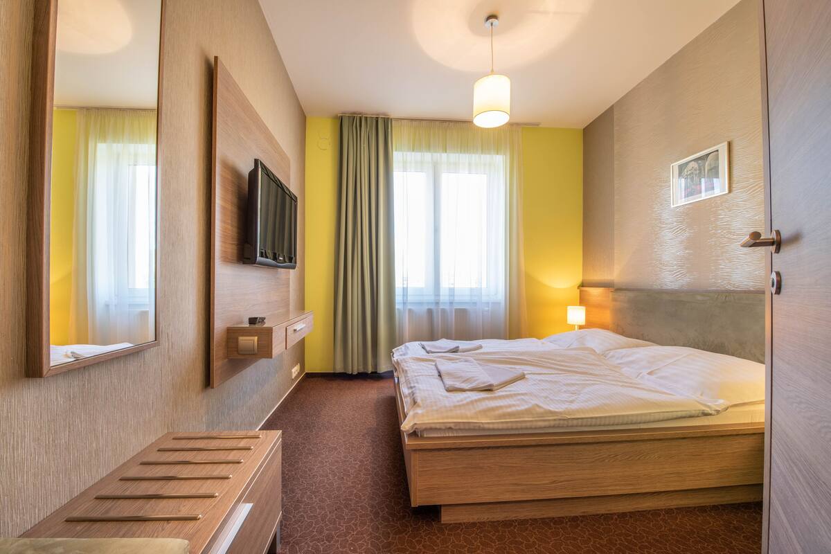 Quarto de hotel com cama, mesa de cabeceira, televisão, janela com cortinas e porta.