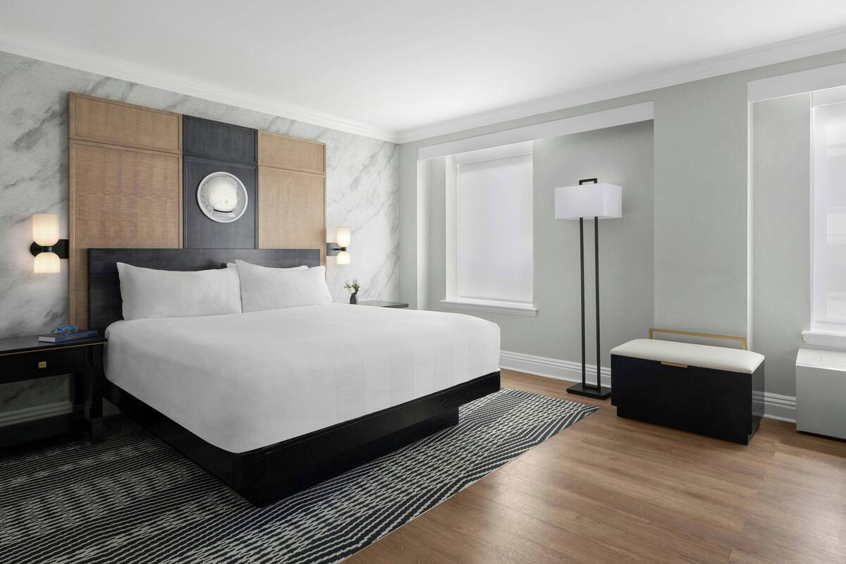 Ein modernes Hotelzimmer mit einem großen Bett, einem Nachttisch, einer Nachttischlampe, einer Stehlampe und einer Bank.