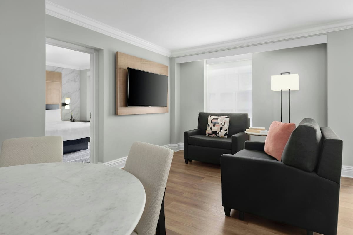 Ein modernes Hotelzimmer mit einem wandmontierten Flachbildfernseher, einem Sofa, einem Esstisch mit Stühlen und einer Stehlampe.