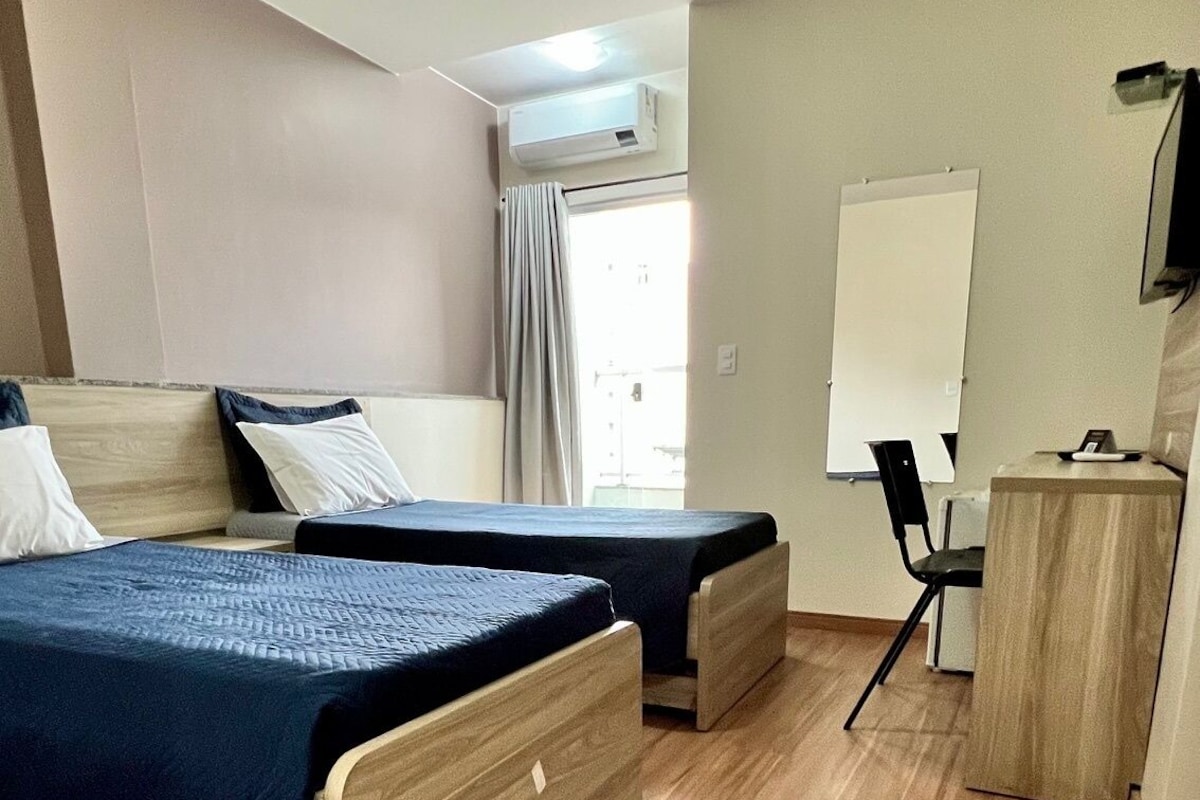 A hotel room with two beds, a desk, a chair, a whiteboard, and an air conditioning unit.