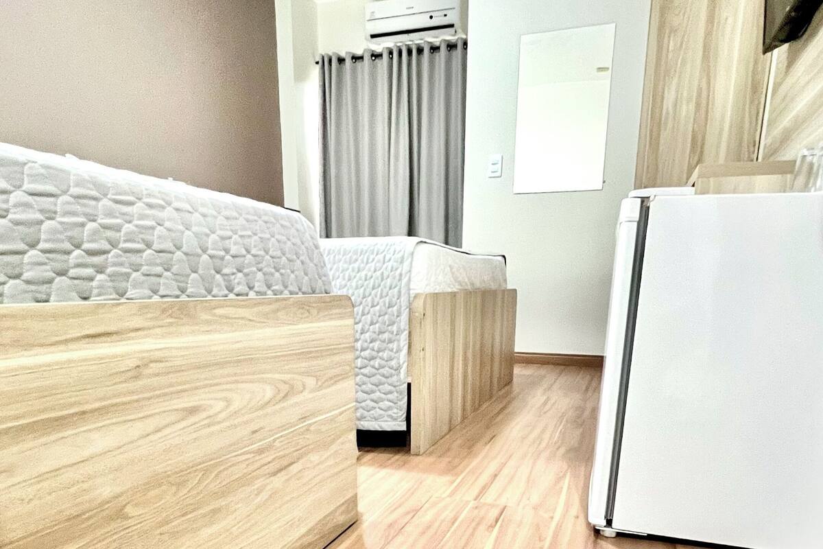 A hotel room with a wooden floor, a bed with a white quilt, a desk, a chair, a refrigerator, and a window with curtains.