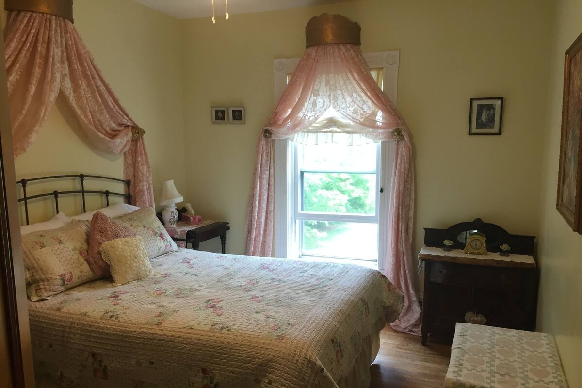Un dormitorio con una cama con dosel, una mesita de noche, un aparador y una ventana con cortinas.