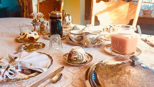 A table set with fine china, silverware, and a coffee pot, indicating a formal dining setting. - Evangeline's Tower Bed & Breakfast (Parrsboro)