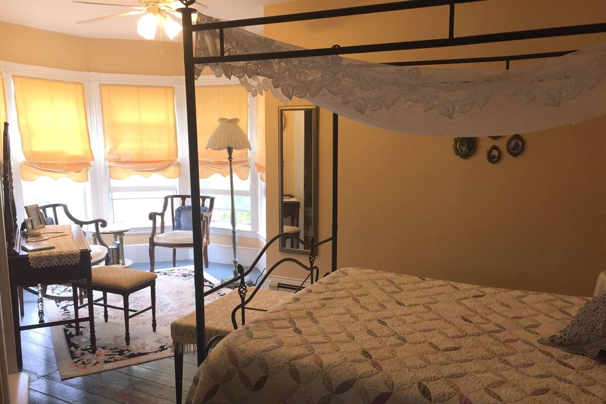 Un dormitorio con cama con dosel, una silla, una mesa y un ventanal con cortinas.