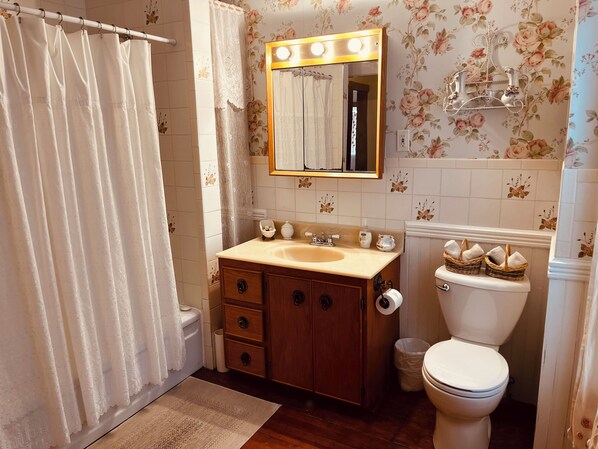 A bathroom with a wooden vanity, a sink, a mirror with lights, a toilet, and a shower curtain. - Evangeline's Tower Bed & Breakfast (Parrsboro)