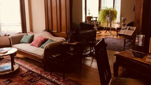 A cozy living room with a sofa, a wooden table, and a rocking chair. There is a large window with curtains, a potted plant, and a fan. - Evangeline's Tower Bed & Breakfast (Parrsboro)