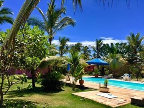 A backyard with a swimming pool, palm trees, and a thatched-roof structure.