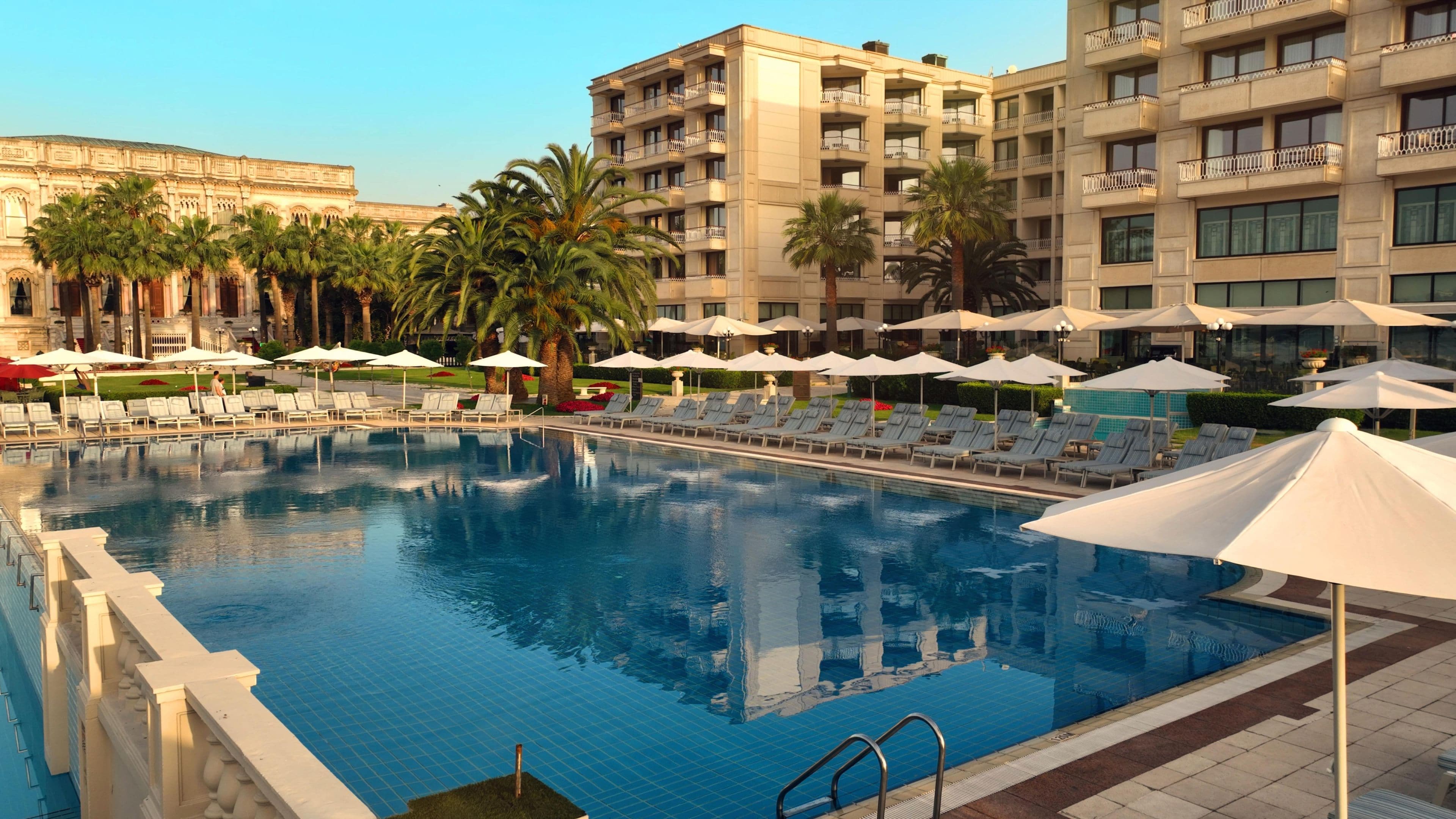 A swimming pool with lounge chairs and umbrellas, surrounded by palm trees and a hotel building.