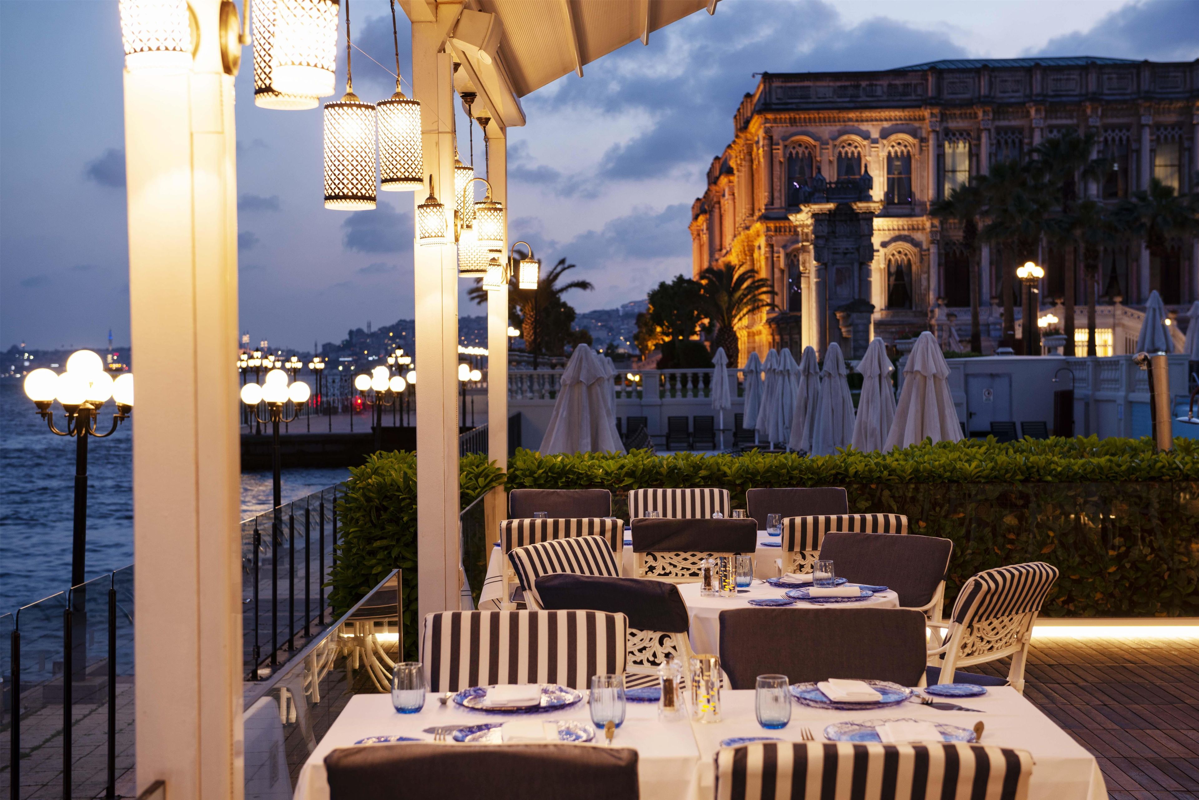 An outdoor dining area with tables and chairs, overlooking a body of water and a historic building in the background.