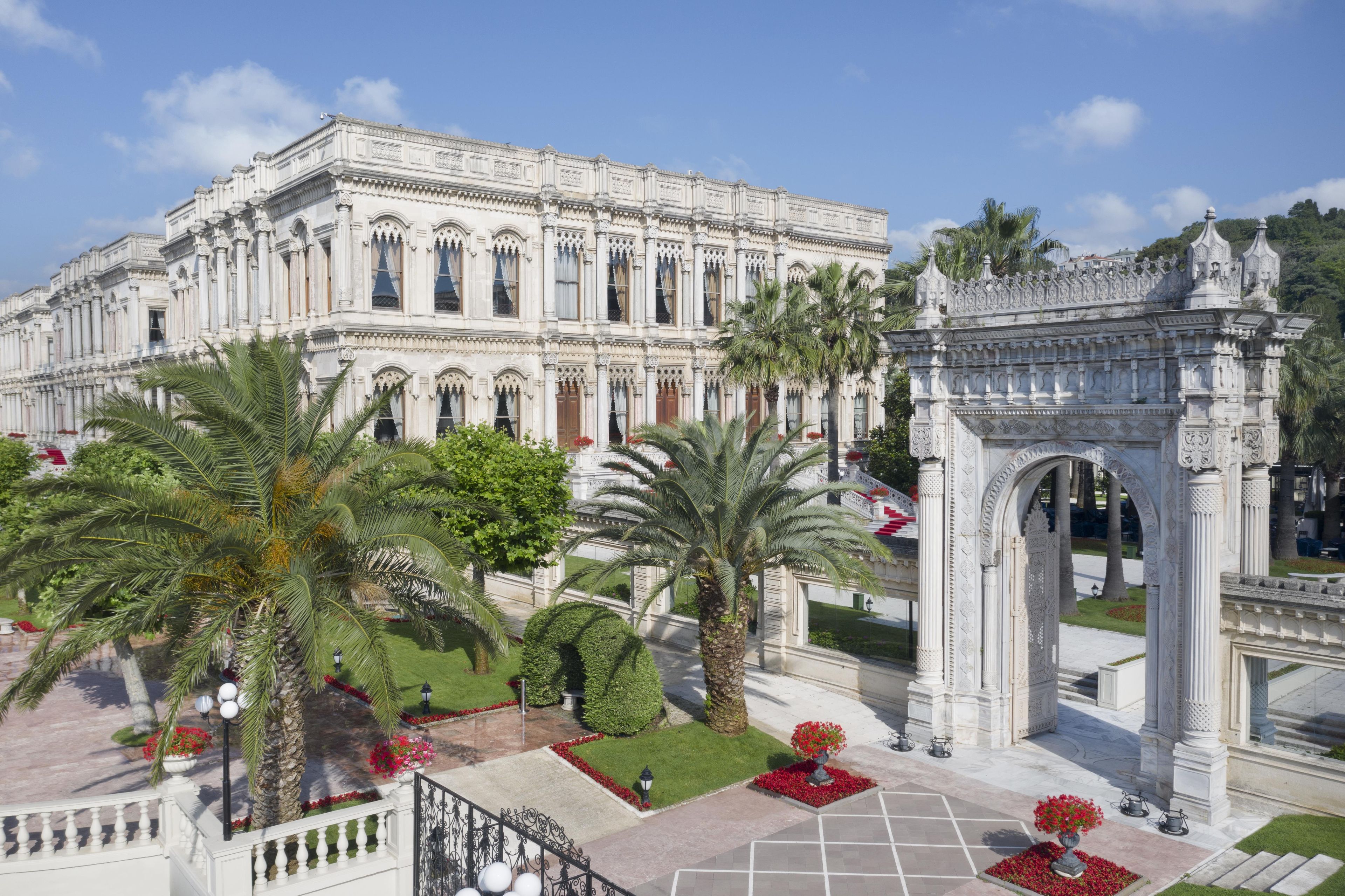 A grand building with ornate architecture, a central archway, and well-maintained gardens with palm trees and red flowers.