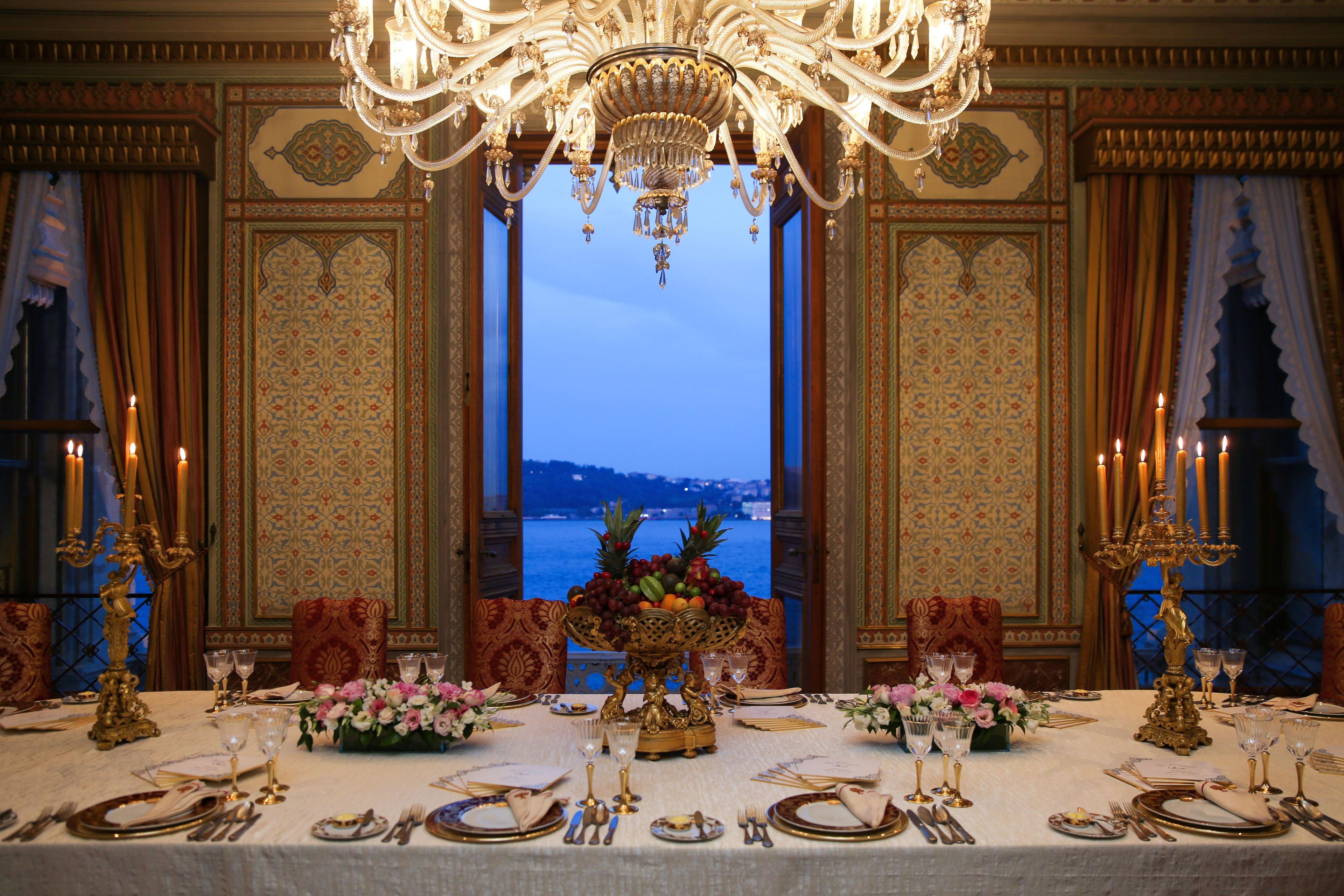 An opulent dining room with a view of a body of water, ornate chandeliers, and a table set for a meal.