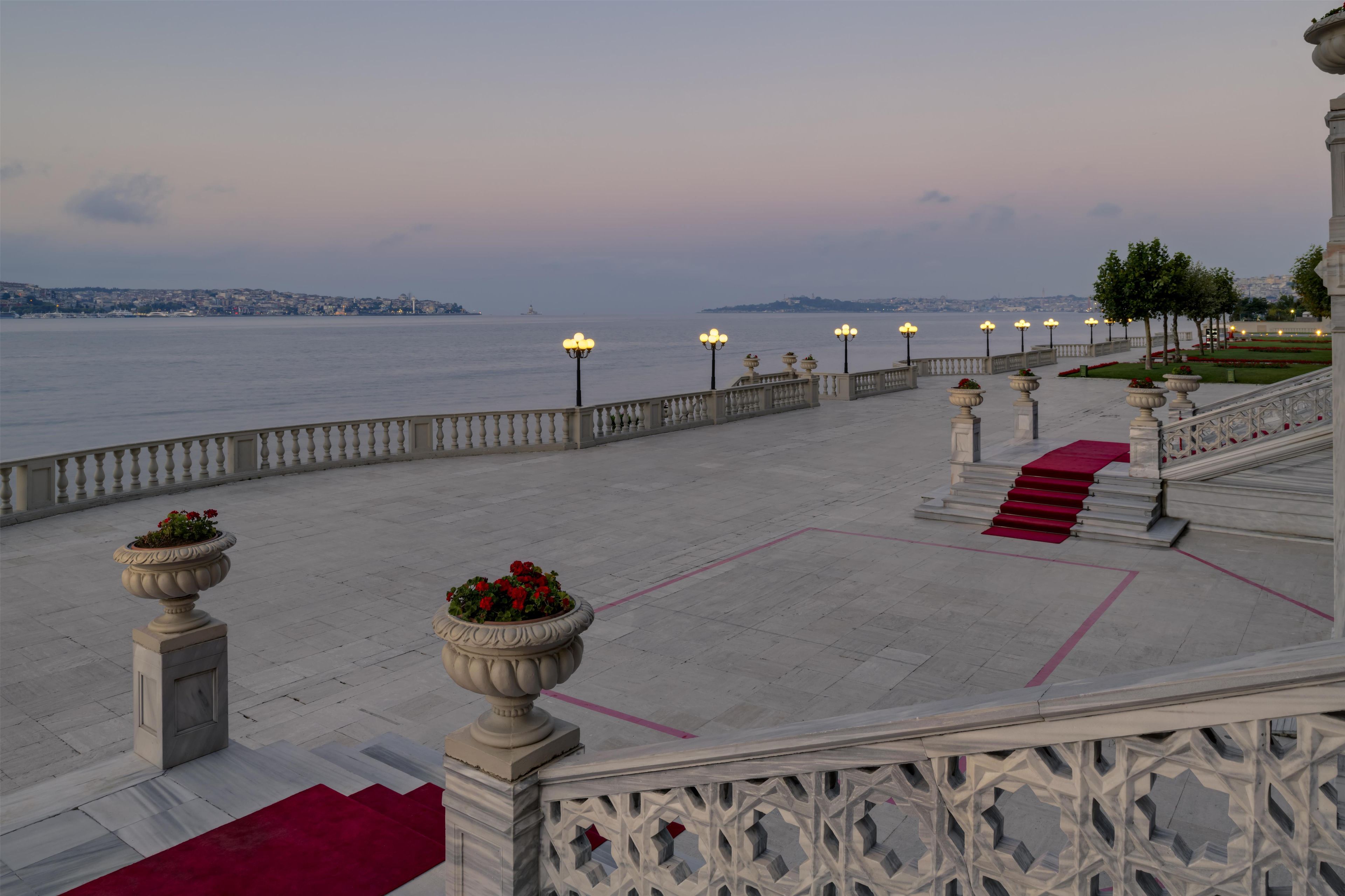 A waterfront promenade with a red carpet leading to a staircase, bordered by balustrades and street lamps.