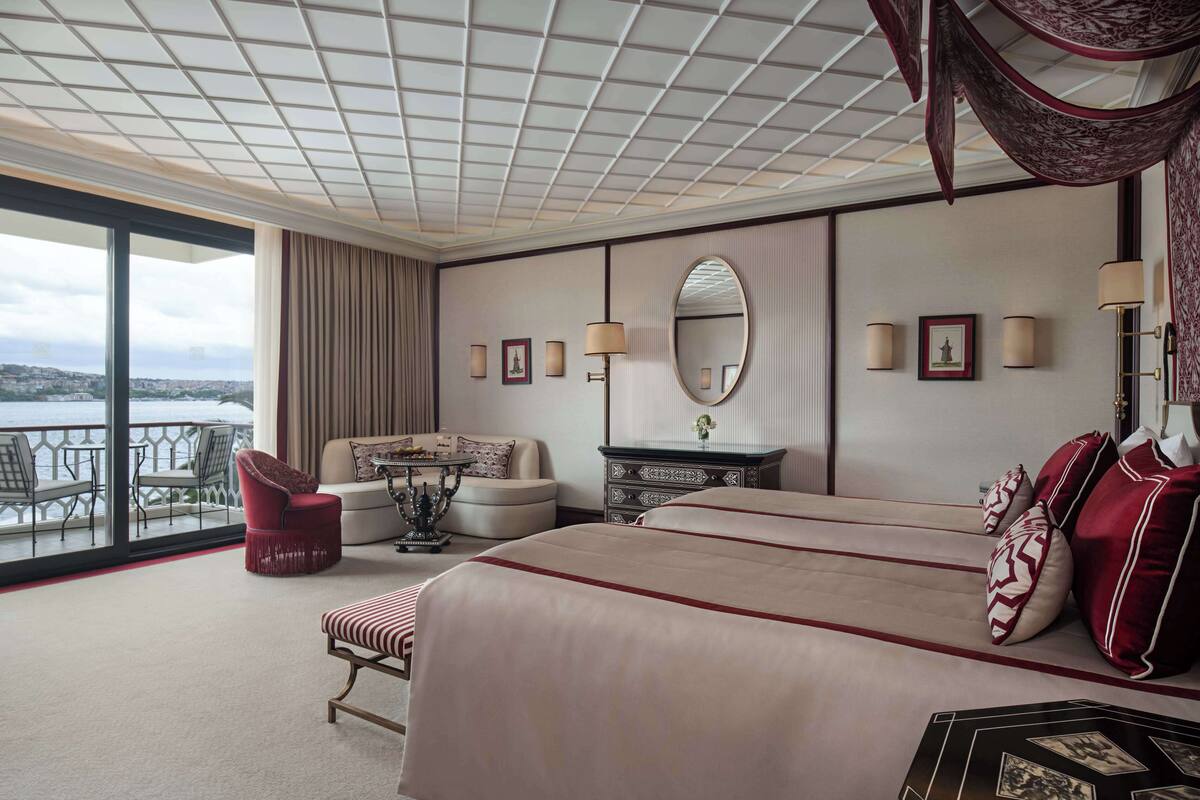 A hotel room with a large bed, a sofa, a red chair, a small table, and a view of the city.