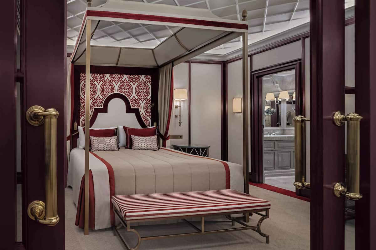 A four-poster bed with a red and white patterned headboard, a bench, and a small table beside it.