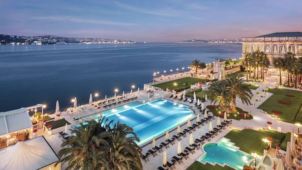 Una amplia piscina al aire libre con tumbonas y sombrillas, rodeada de césped cuidado y palmeras, con vista al mar y un edificio histórico al fondo.