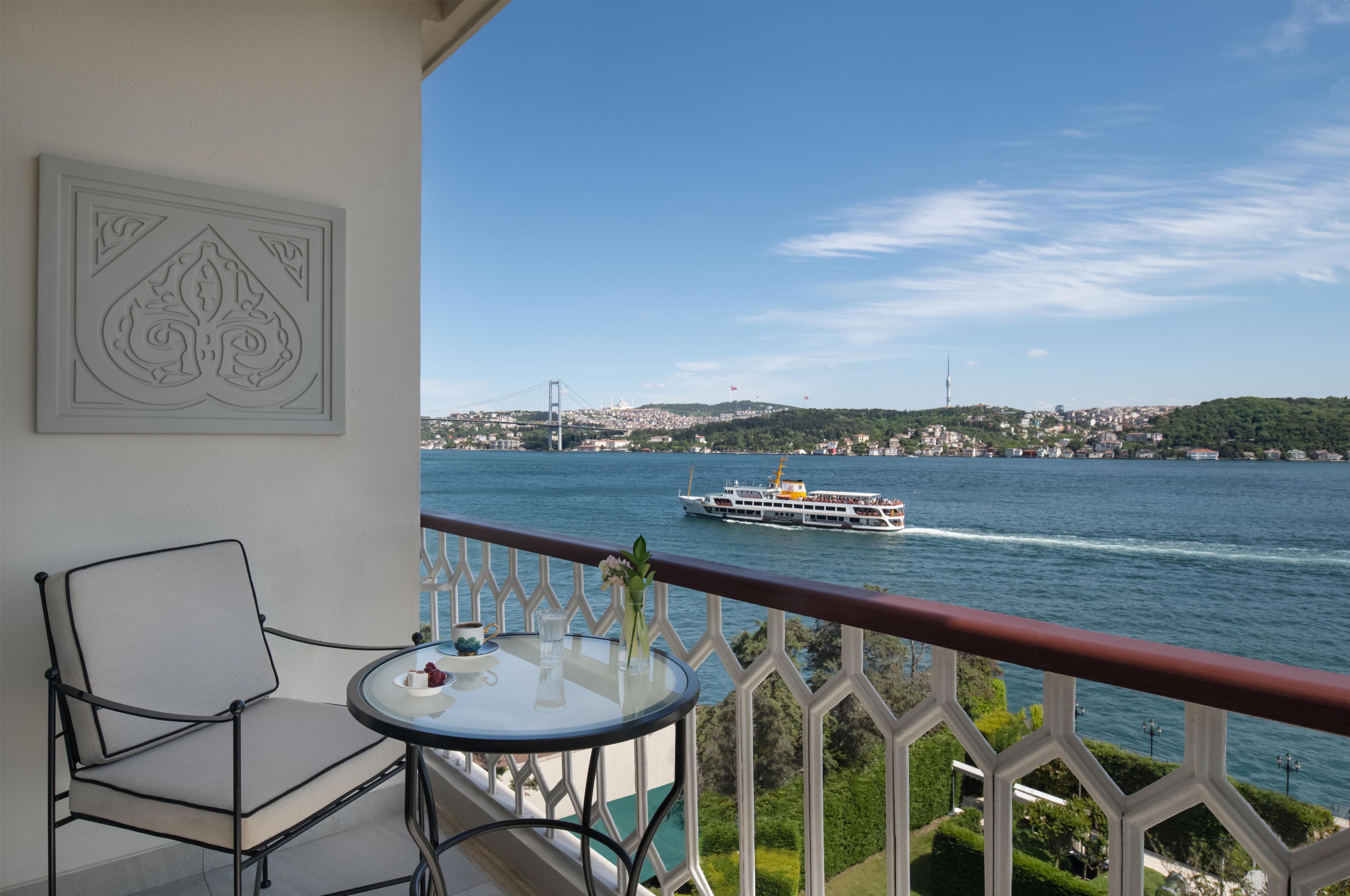A balcony with a table and chair, overlooking a river with a boat and distant cityscape.