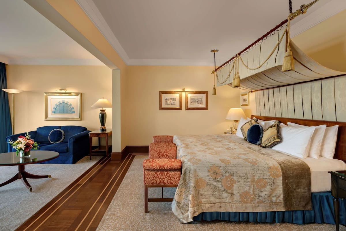 A hotel room with a bed, a sofa, a chair, a table with flowers, and framed pictures on the wall.