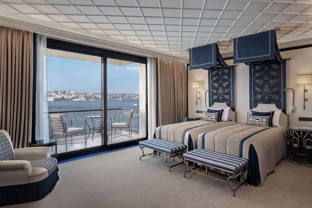 A hotel room with a large balcony, a bed with matching headboard and footboard, a bedside table, and a chair.