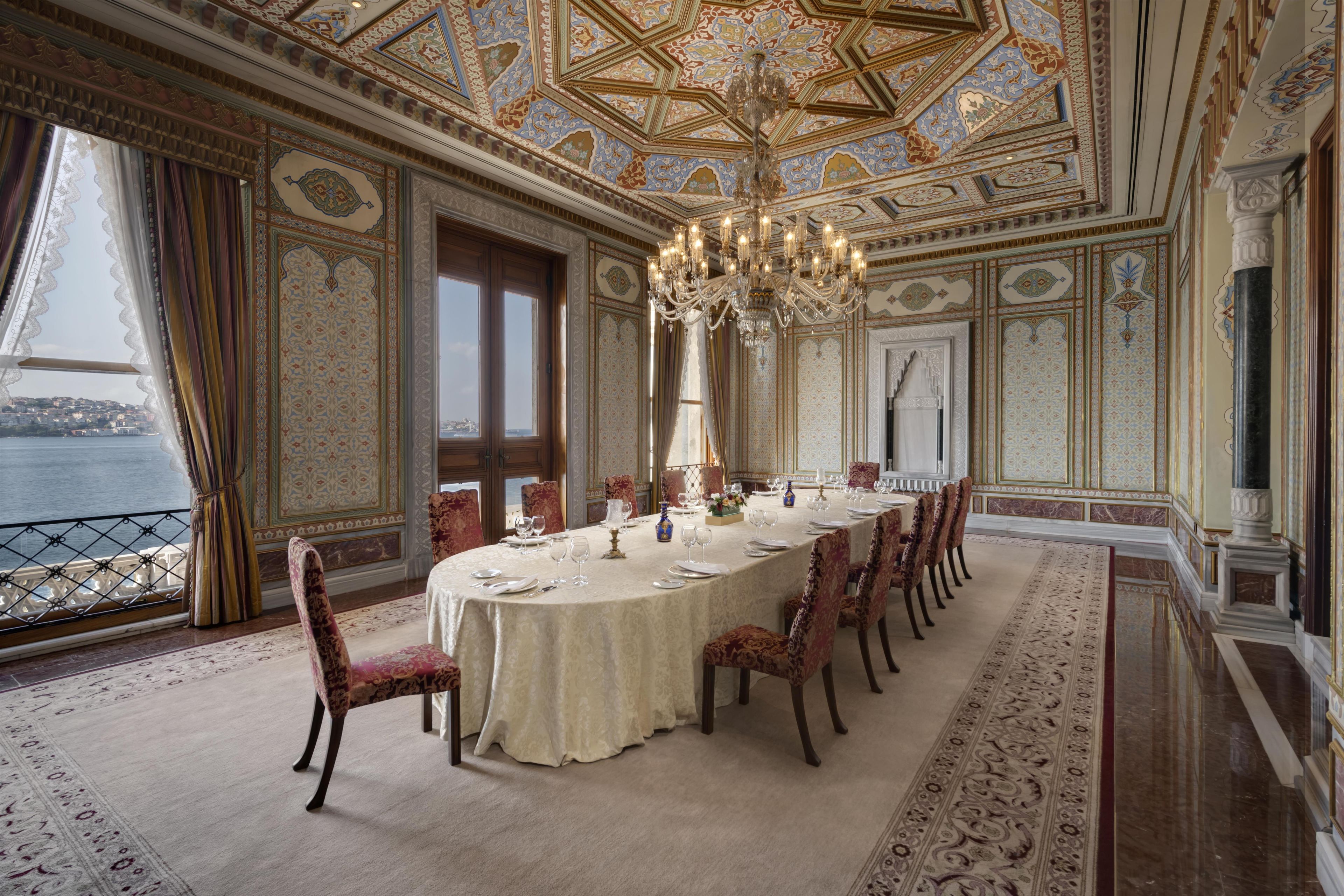 Een statige eetzaal met een grote ronde tafel, versierde stoelen en een kroonluchter.