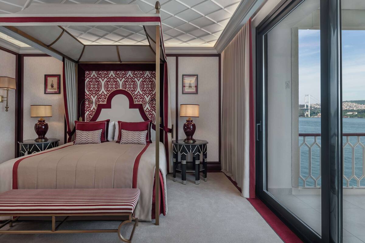 A four-poster bed with a canopy, two bedside tables with lamps, a view of a bridge and water, and a balcony with a railing.