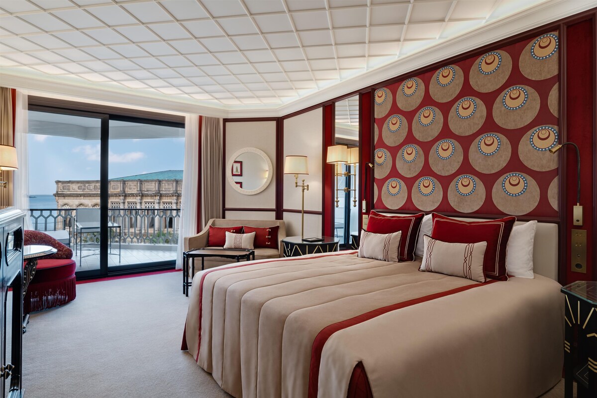 A hotel room with a large bed, a desk, a chair, a balcony with a view of a building, and a patterned wall.