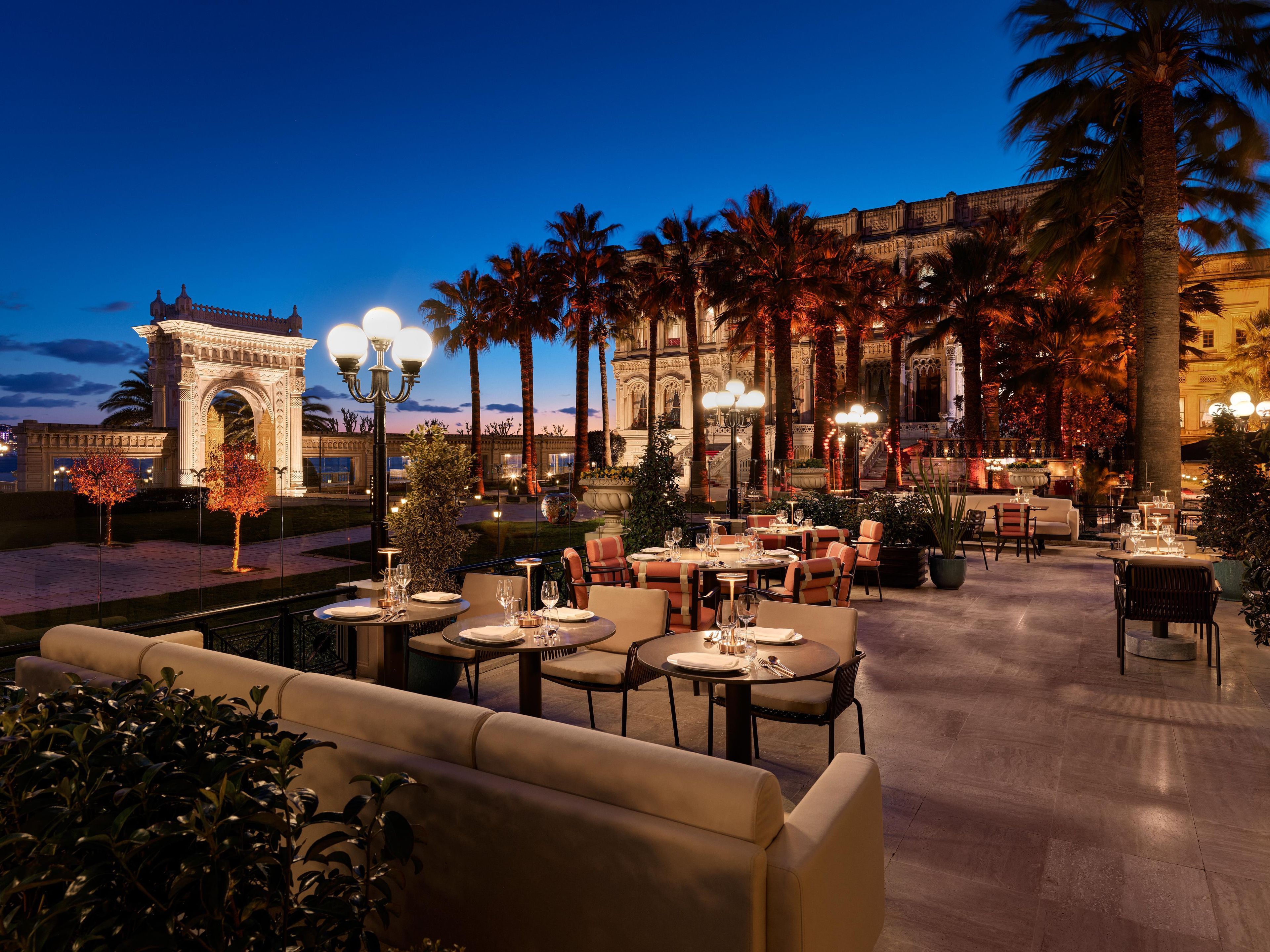 An outdoor dining area with tables and chairs, palm trees, and a building with an ornate archway in the background.