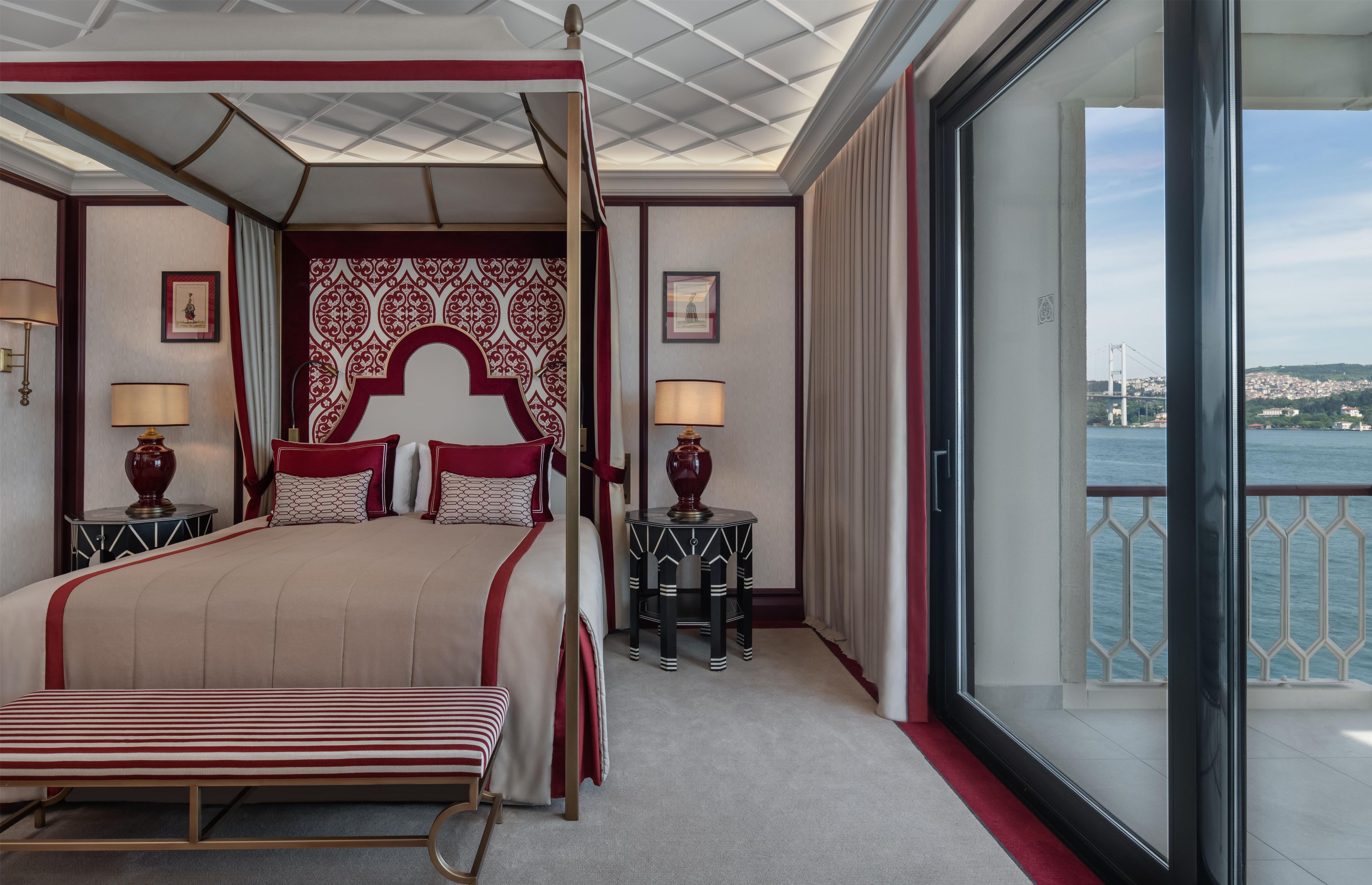Una cama con dosel y cortinas, dos mesitas de noche con lámparas, un ventanal con vistas a un puente y al paisaje urbano, y una pared con diseño.