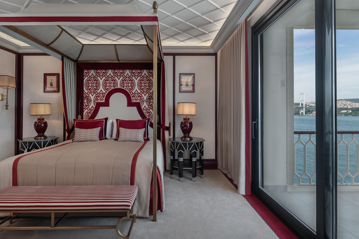 Una cama con dosel y cortinas, dos mesitas de noche con lámparas, un ventanal con vistas a un puente y al paisaje urbano, y una pared con diseño.