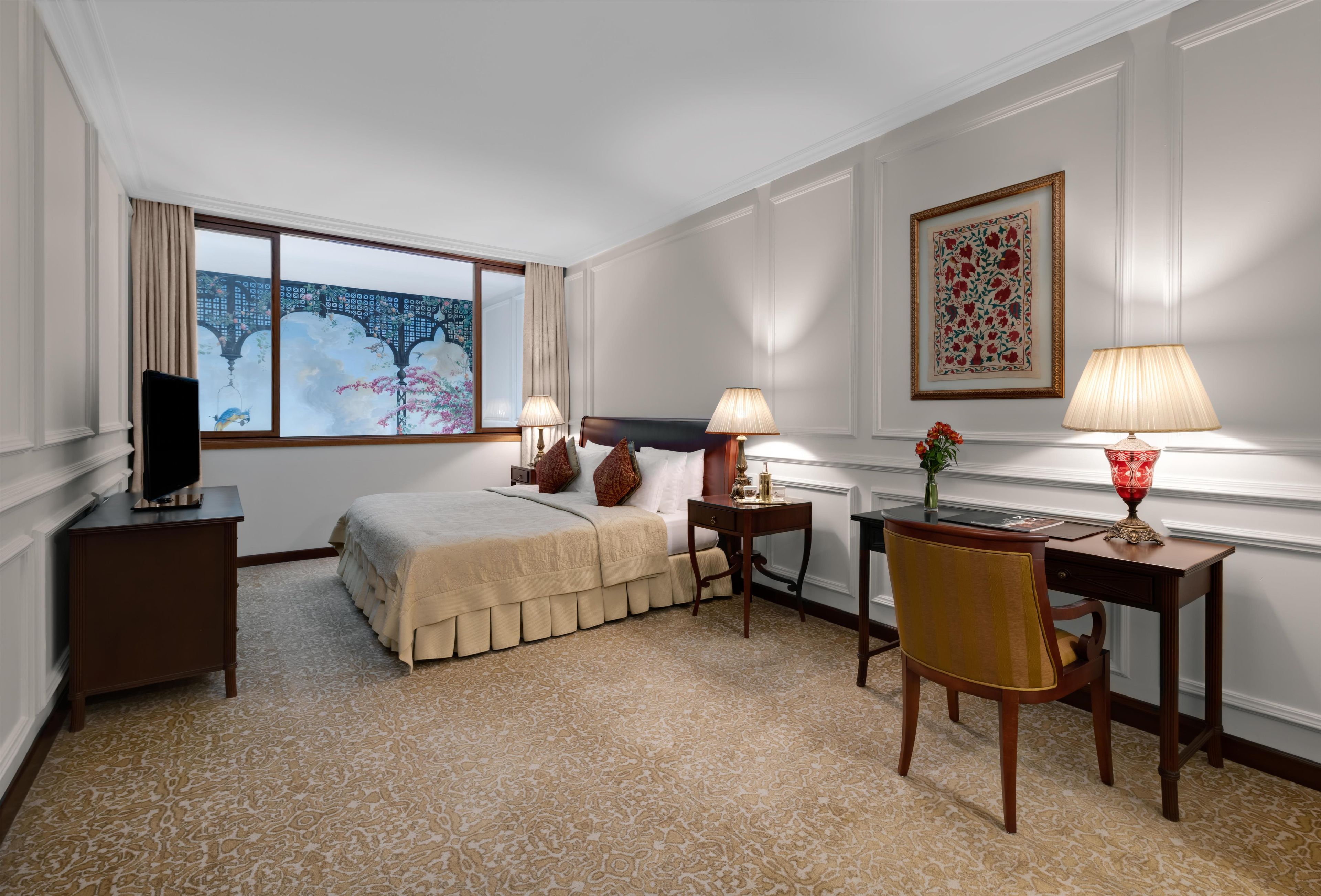 Habitación de hotel con cama, dos mesitas de noche, un escritorio con silla, televisión y un ventanal con vistas a un edificio.