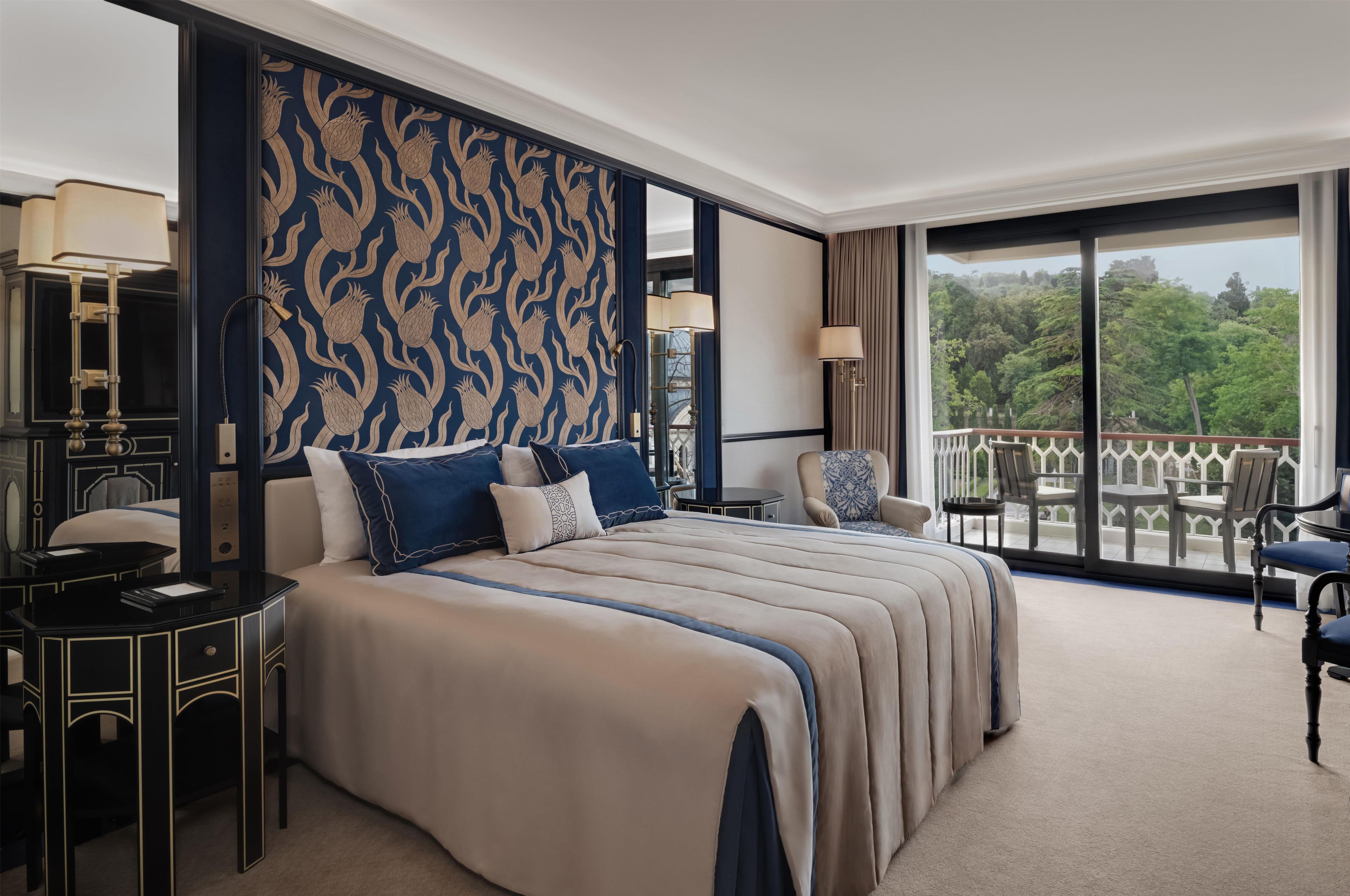 Habitación de hotel con una cama grande, dos mesitas de noche, un área de descanso con una silla y un balcón con vista a árboles.