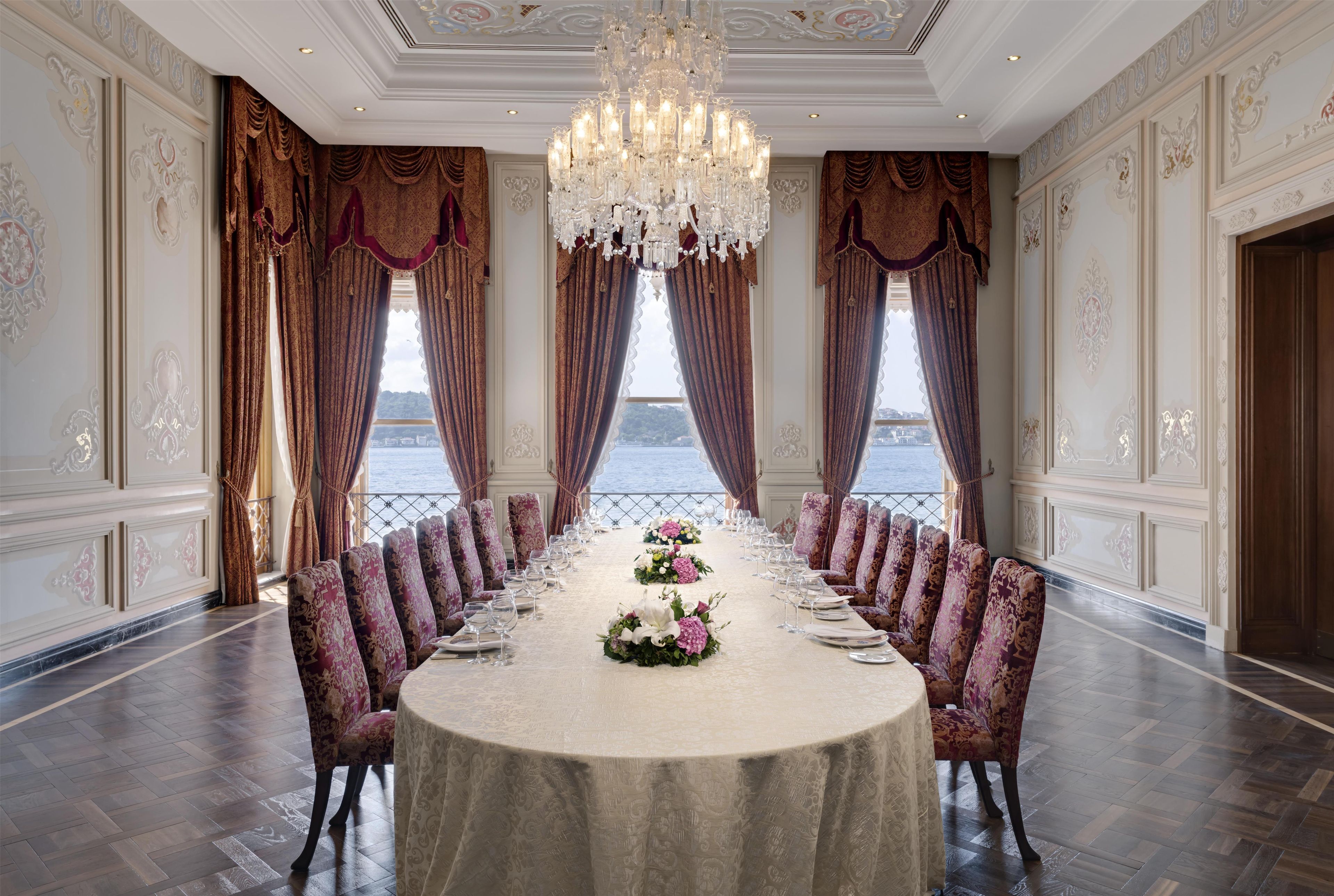 A formal dining room with a long table set for a meal, large windows with curtains, and a chandelier.