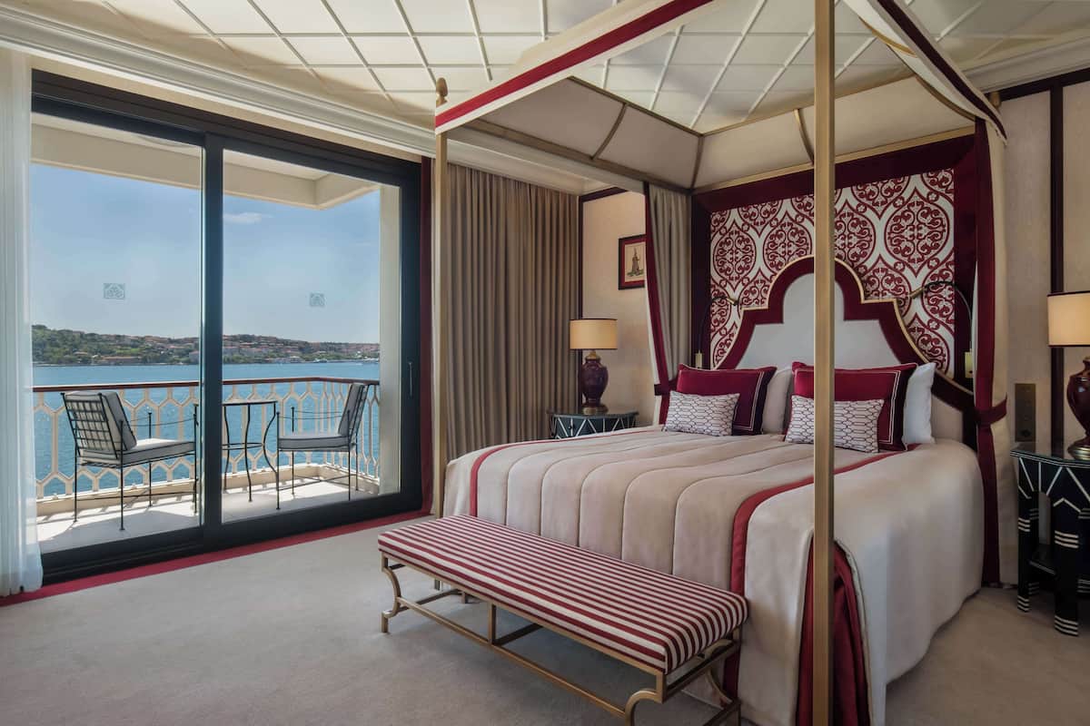 A four-poster bed with a view of the sea, a balcony with chairs, and a large window.