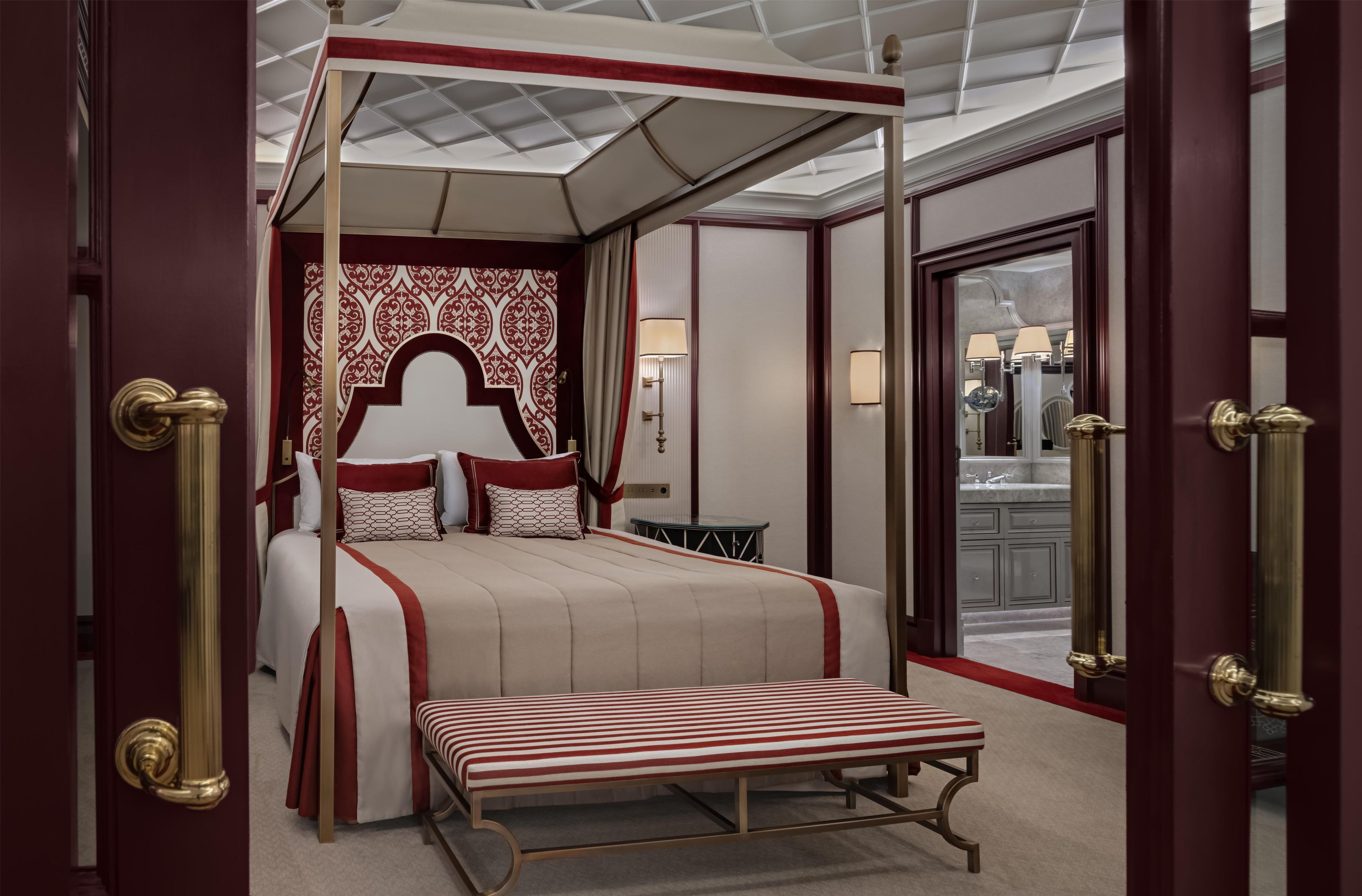 A four-poster canopy bed with a red and white color scheme, a bench, and a view of a bathroom with a mirror and a sink.