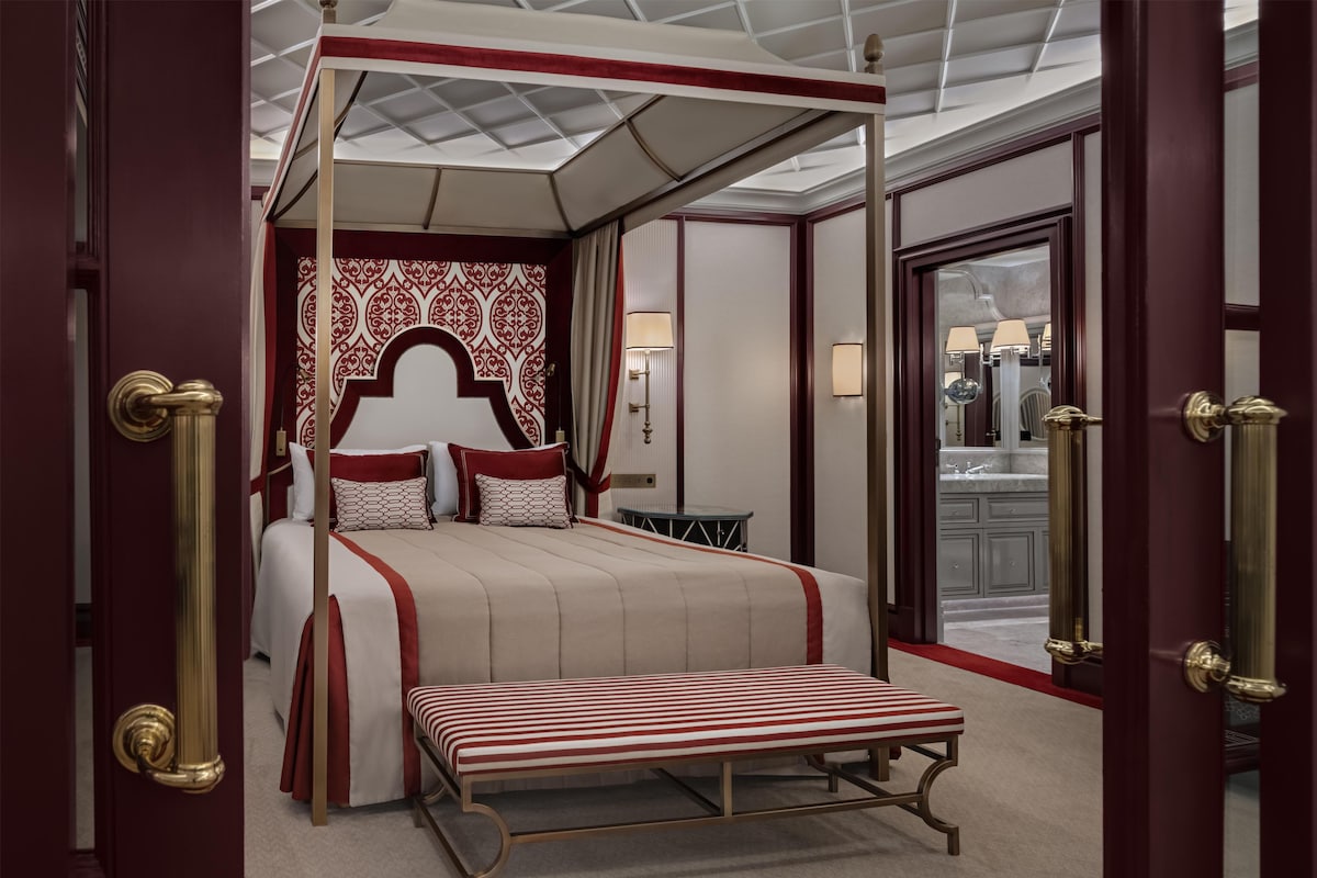 Una cama con dosel y cuatro columnas, con una decoración en blanco y rojo, un banco y vistas a un baño con espejo y lavamanos.