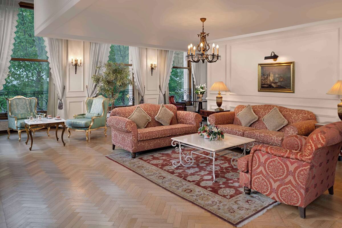 A spacious living room with a large sofa set, a coffee table, and a chandelier. There are two armchairs, a side table, and a potted plant.